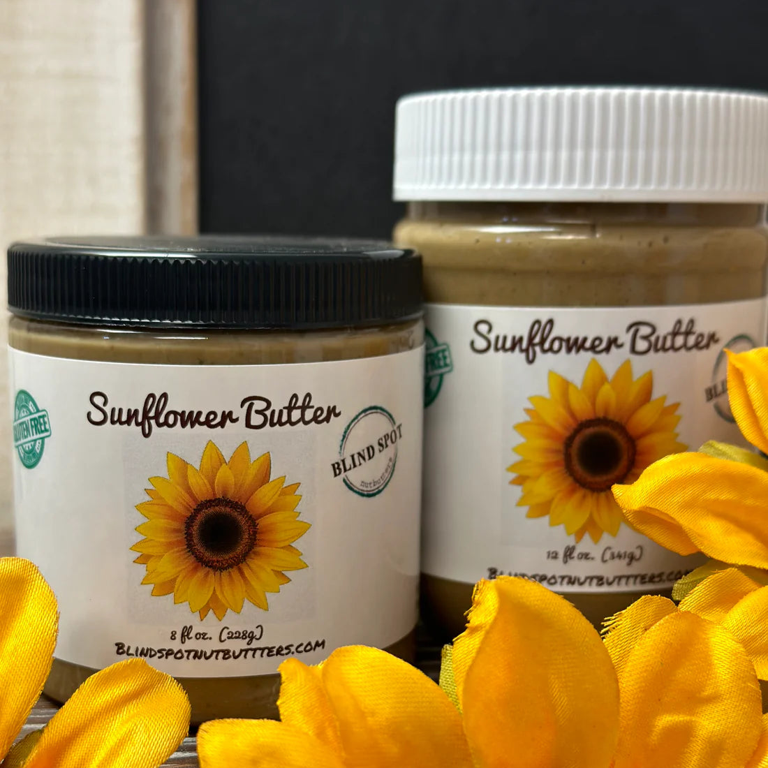 Two jars of Sunflower Seed Butter 🌻 – Pure, Roasted, and Seriously Good sit side by side with sunflower labels; one has a black lid, the other white. Yellow sunflower petals are in front. This nut-free spread contains no added sugar.