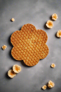 Thumbnail for A Flower-Shaped Summer Gram Original Stroopie Single – Classic Caramel sits on a gray surface, surrounded by small, light yellow flower decorations, hinting at its gooey caramel center and springtime fun.