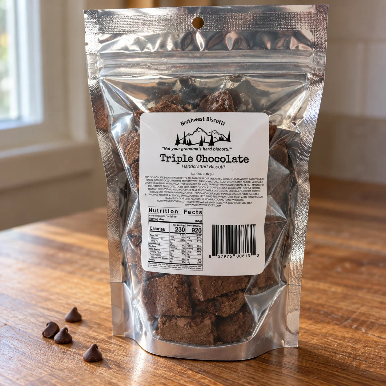 A stand-up resealable pouch of Northwest Biscotti Triple Chocolate Handcrafted Biscotti cookies on a wooden table, with a few chocolate chips placed in front and a softly blurred indoor background.