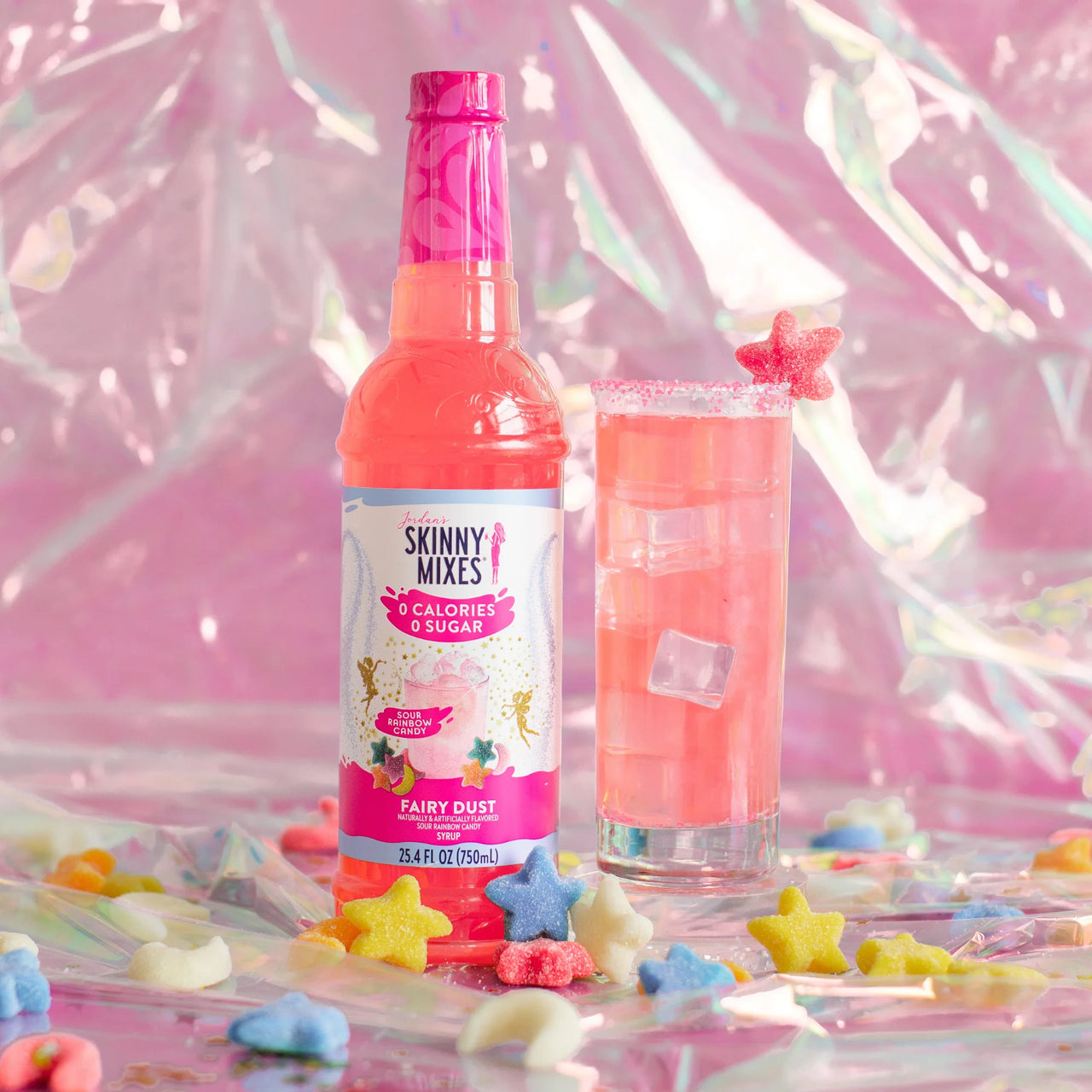 A pink bottle of Fairy Dust Skinny Syrup – Sweet, Sour & Totally Spellbinding 🧚‍♀️🍬✨ stands beside a glass of pink iced drink with a star garnish. Colorful star candies decorate the pastel, iridescent background for a keto-friendly touch.