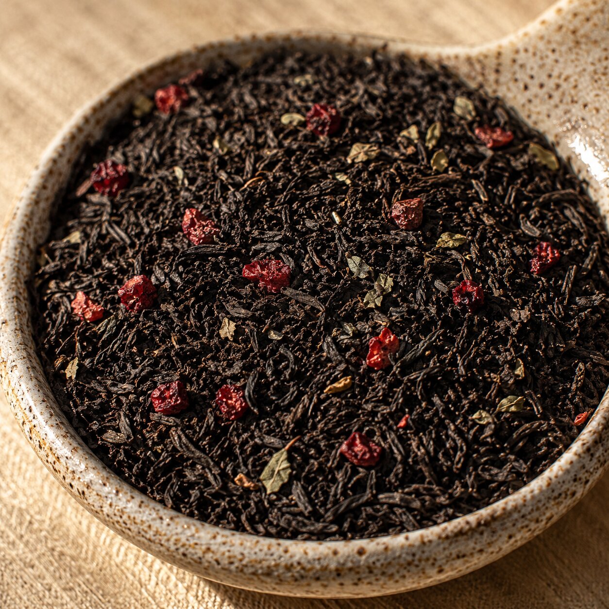 Close-up of black tea leaves with red berries in a ceramic bowl on a wooden surface.