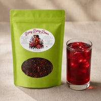 Thumbnail for Green tea package labeled 'Berry Berry Boom' next to a glass of iced berry tea on a beige background