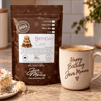 Thumbnail for A bag of Java Momma 'Birthday Cake' whole bean coffee stands on a countertop beside a ceramic mug printed with 'Happy Birthday Java Momma' and a slice of cake on a plate. The scene is set in a real kitchen environment with warm natural light and a blurred plant in the background.