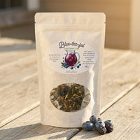 Thumbnail for Tea package labeled 'Blue-tea-ful' with a clear window showing tea leaves, placed on a wooden surface with blueberries beside it.