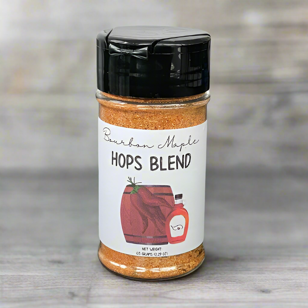 A jar of Bourbon Maple Hops Seasoning 🥃 rests on a gray wooden surface—ideal as a smoky chipotle grilling rub to add rich depth to your dishes.