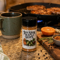 Thumbnail for Bourbon Maple Hops Seasoning bottle in a cozy kitchen setting with grilling in the background.