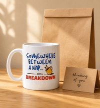 Thumbnail for White mug with humorous text Somewhere between a nap and a breakdown 
design, brown paper bag, and card on a wooden surface.