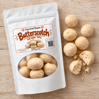 Thumbnail for A white stand-up pouch labeled 'Butterscotch Salt Water Taffy' with a clear window showing round beige taffy balls, placed on a light wood surface beside several loose taffy pieces, one of which is broken open to show the interior texture.