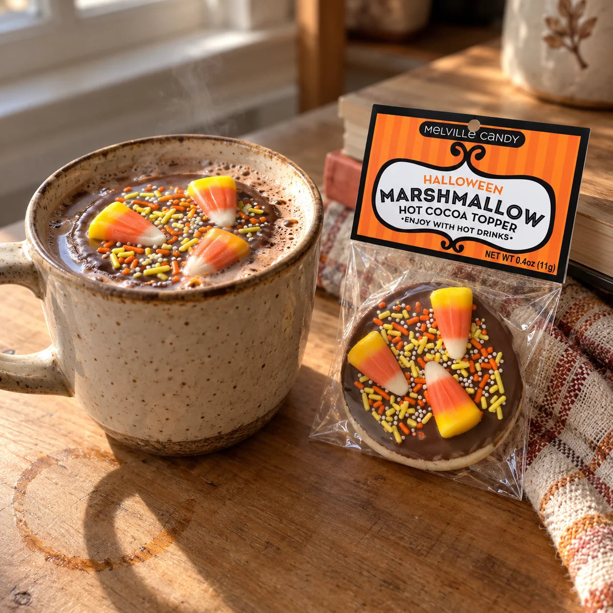 A packaged marshmallow labeled Melville Candy Halloween Marshmallow Hot Cocoa Topper sits beside a ceramic mug filled with hot cocoa and candy corn sprinkles on a wooden table.
