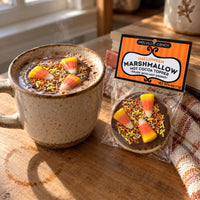 Thumbnail for A packaged marshmallow labeled Melville Candy Halloween Marshmallow Hot Cocoa Topper sits beside a ceramic mug filled with hot cocoa and candy corn sprinkles on a wooden table.