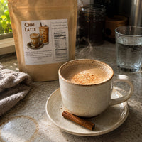 Thumbnail for A ceramic cup of steaming chai latte sits on a saucer with a cinnamon stick, with a stand-up pouch of Chai Latte behind it on a kitchen counter.