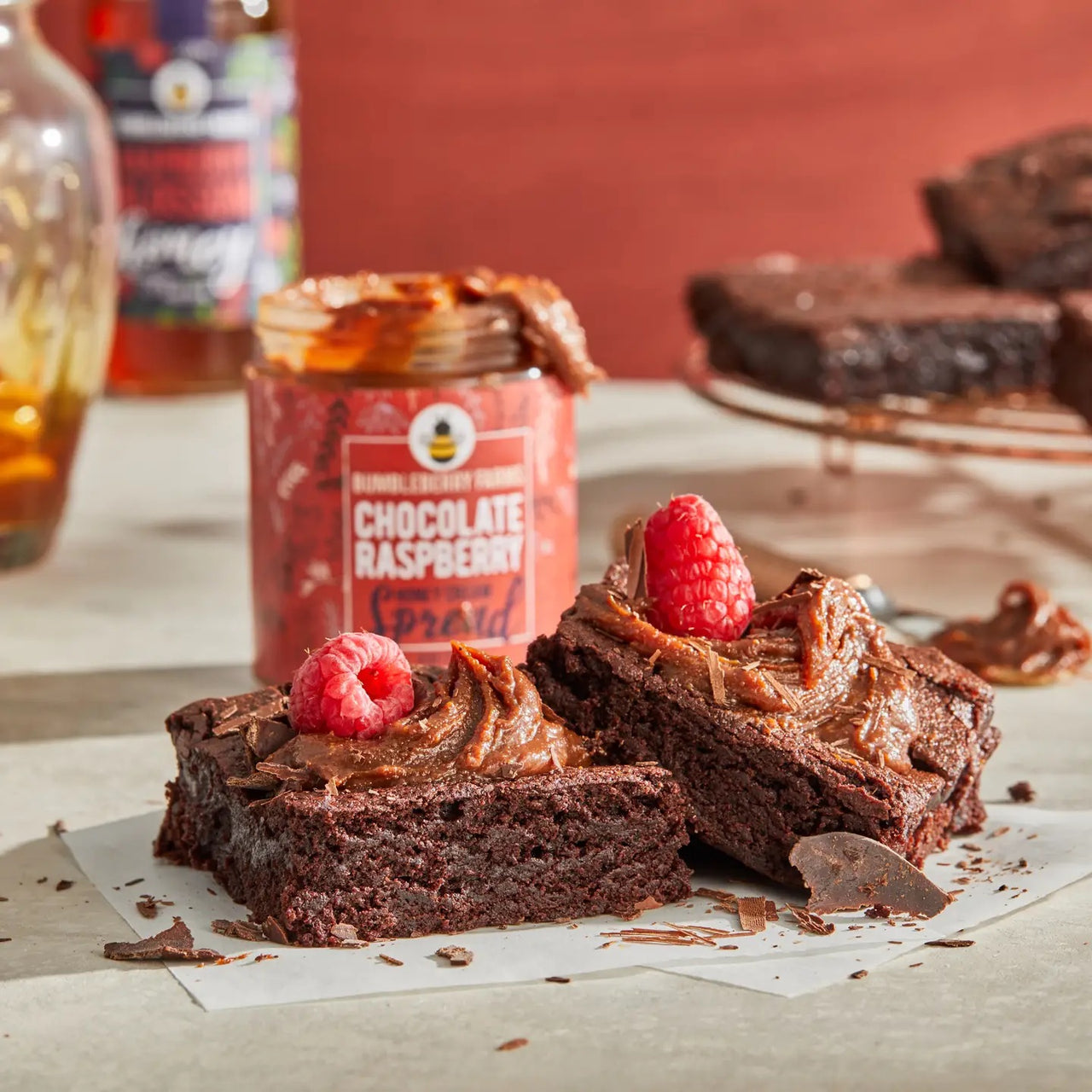Two rich brownies topped with Chocolate Raspberry Honey Cream Spread 🍫 – Award-Winning, Rich & Fruity Chocolate Bliss and a fresh raspberry sit on parchment. An open jar of this spread and extra brownies are seen in the background.