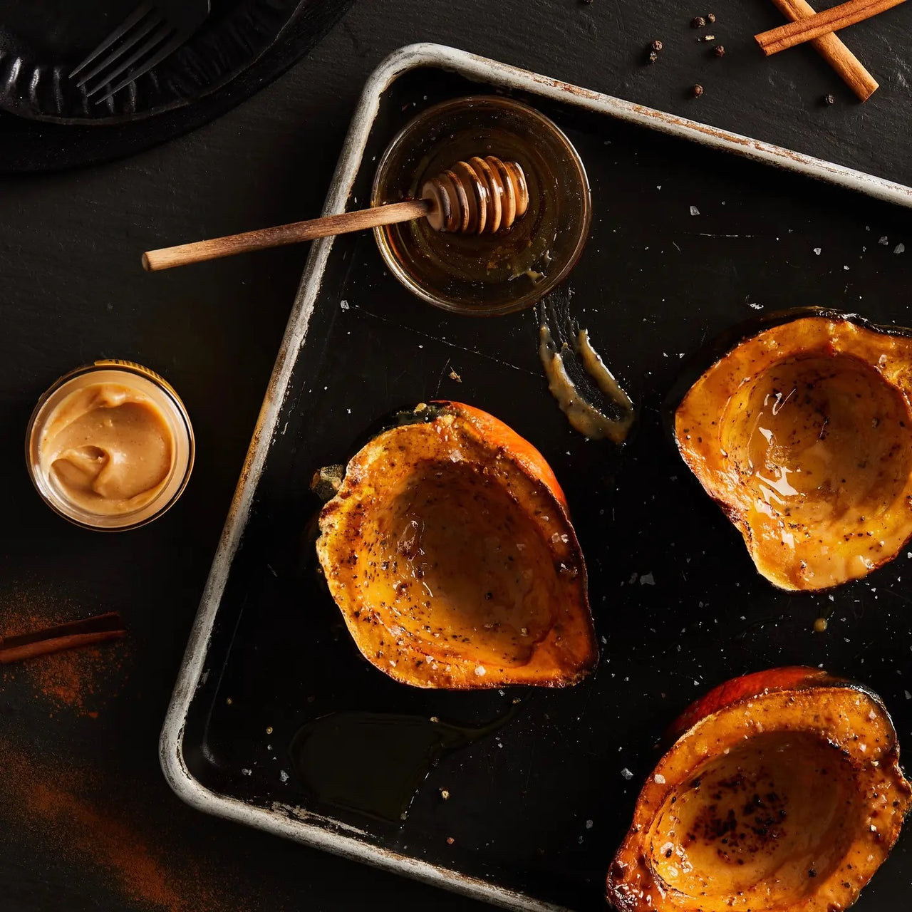 Roasted acorn squash halves on a baking sheet, drizzled with Cinnamon Stick Honey Cream Spread 🍯 – Cozy Cinnamon Bun Bliss in a Jar. A swirl-topped jar, honey dipper, cinnamon sticks, and peppercorns sit nearby on a dark surface.