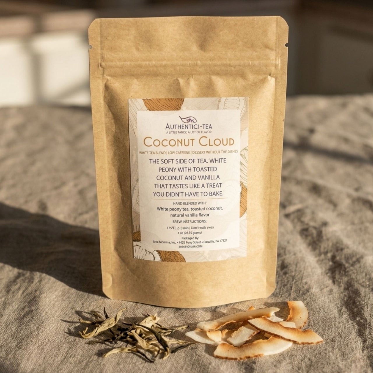 Coconut Cloud Authentici-tea white tea bag standing upright on gray linen with scattered white peony leaves and toasted coconut flakes