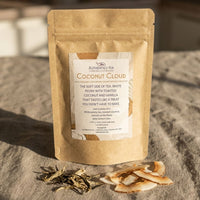 Thumbnail for Coconut Cloud Authentici-tea white tea bag standing upright on gray linen with scattered white peony leaves and toasted coconut flakes
