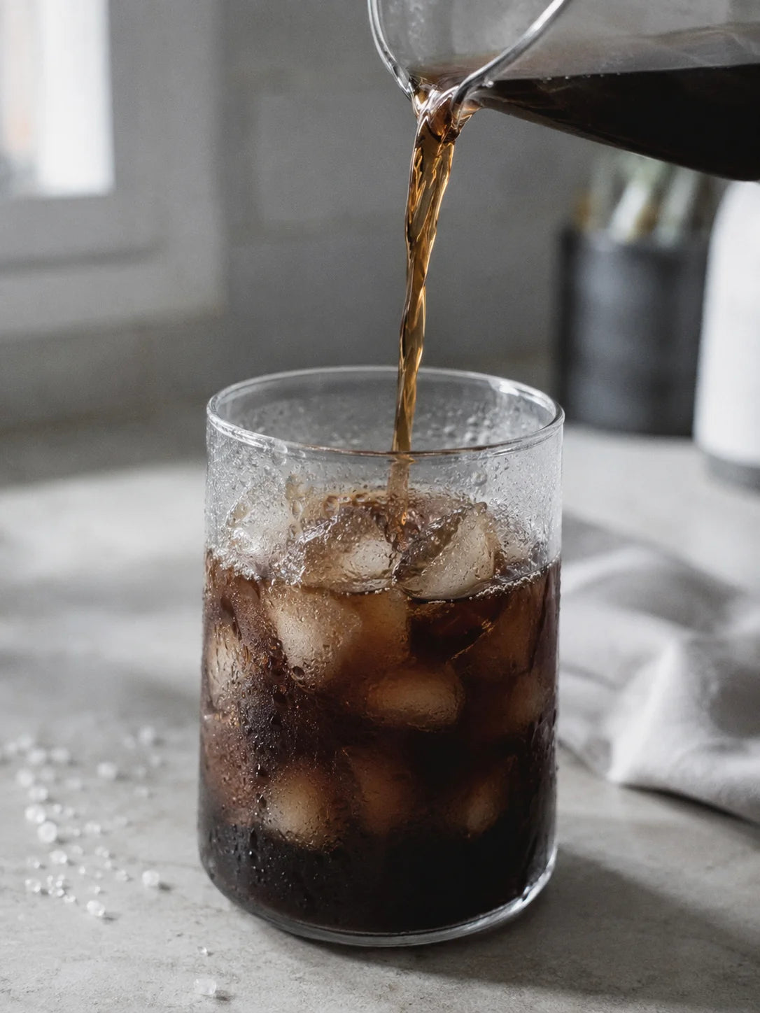 Pouring iced coffee into a glass with ice cubes on a light surface.