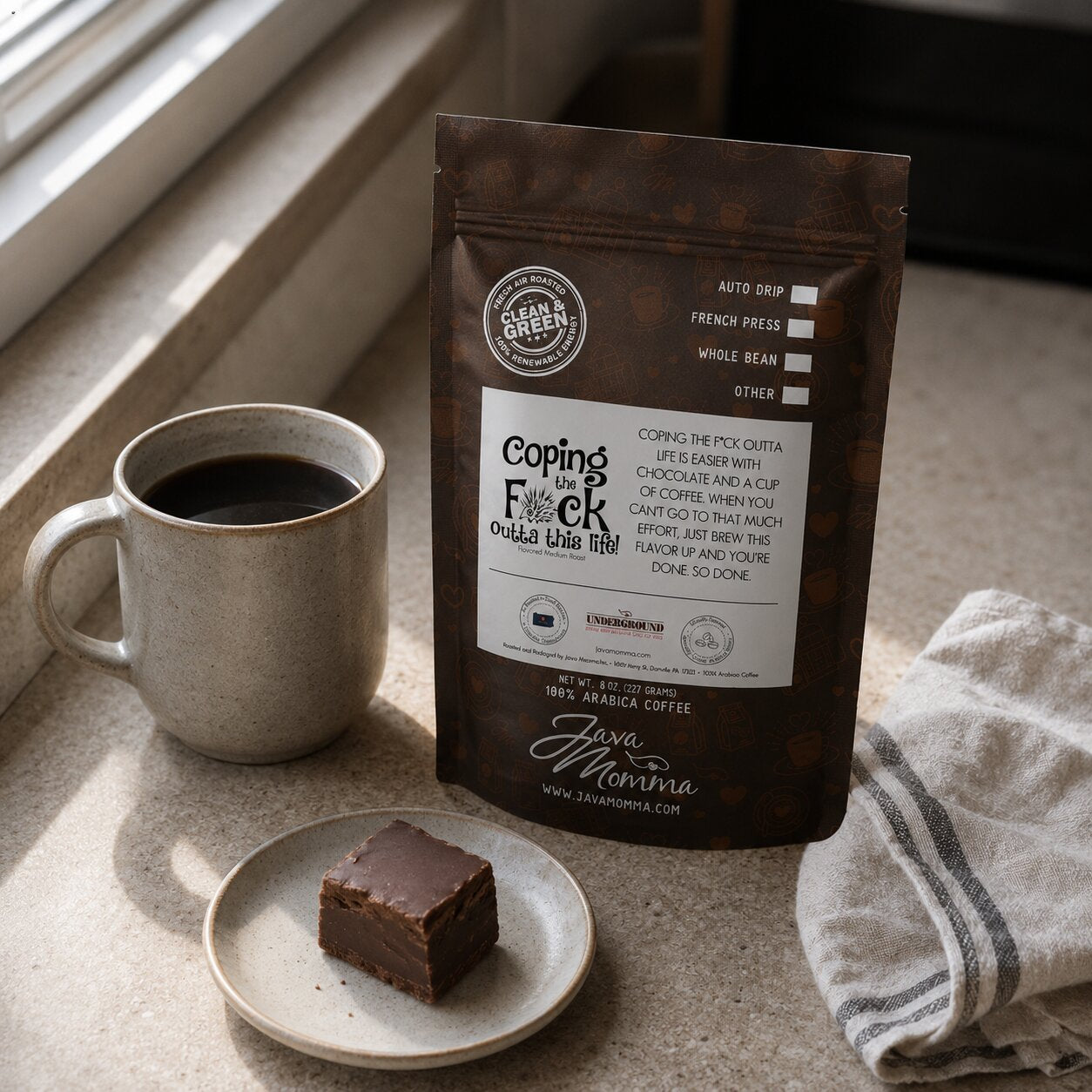 Java Momma Coping the F%k Outta This Life coffee bag next to a mug of coffee with a piece of chocolate fudge on a small plate