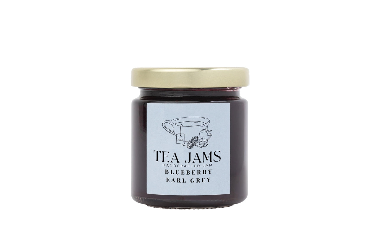 A glass jar from the Mini Tea Jams Gift Set 🍯 contains handcrafted Blueberry Earl Grey tea-infused jam with a gold lid and label featuring a teacup, blueberries, and tea leaves—flavors inspired by Harney & Sons teas.