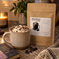 Thumbnail for Dairy Free Dark Chocolate Sea Salt Hot Cocoa Mix in a cozy setting with a mug topped with whipped cream.