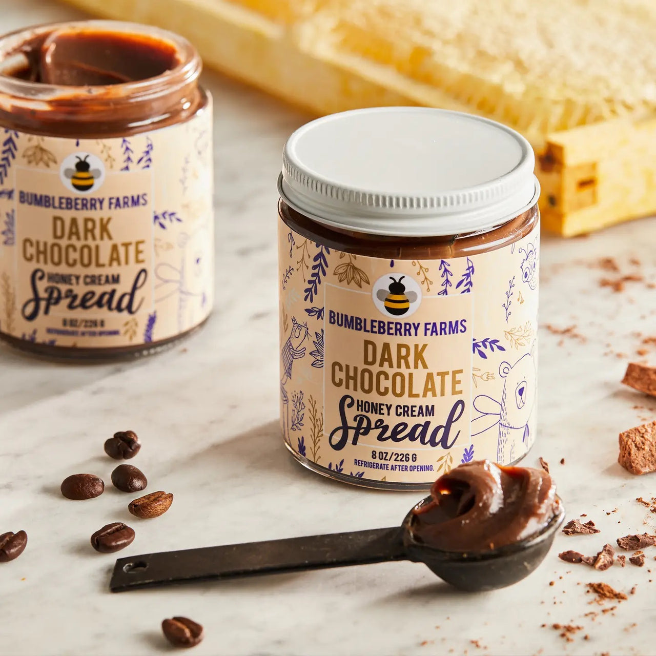 A jar of Dark Chocolate 🍫 Honey Cream Spread – Rich, Spicy, & Decadently Bold sits on a marble surface with a measuring spoon of spread, coffee beans, and honeycomb in the background. Another jar is partially visible.