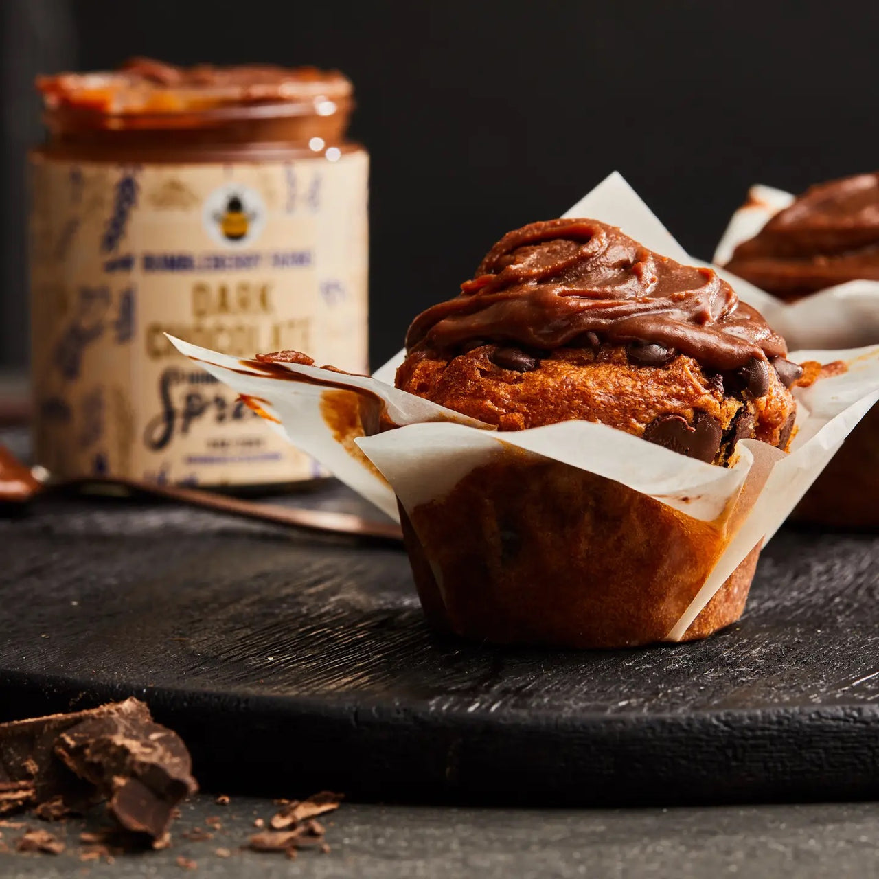 A chocolate muffin with swirled chocolate frosting sits in a white wrapper on a dark surface, accompanied by a jar of Dark Chocolate 🍫 Honey Cream Spread – Rich, Spicy, & Decadently Bold, and pieces of chocolate in the background.