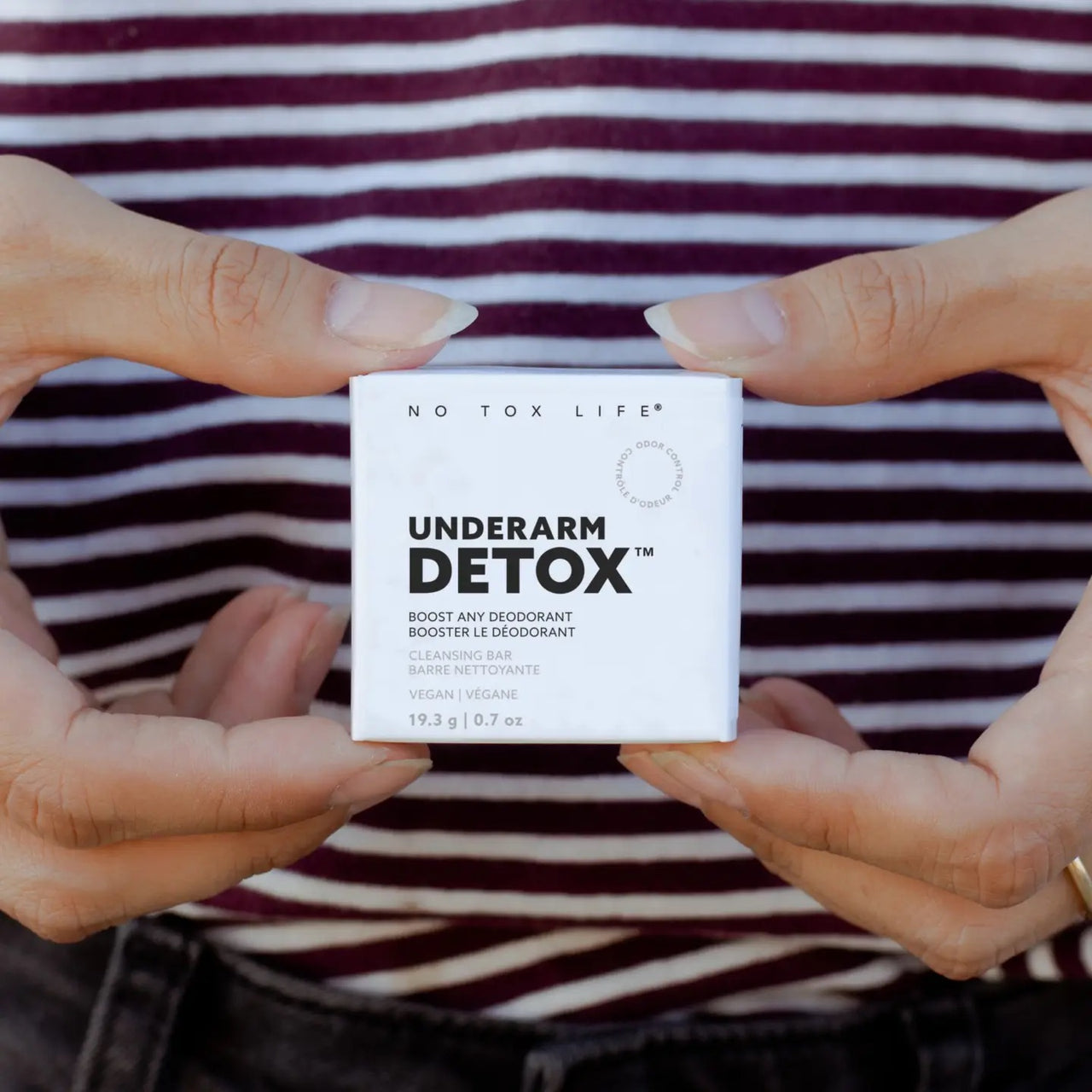 Close-up of hands holding a small white box labeled No Tox Life Underarm Detox – Deodorant Booster Cleansing Bar (19.3g) in front of a person in a maroon and white striped shirt. Ideal for those switching to natural deodorant.