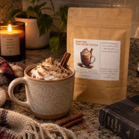 Thumbnail for Dairy Free Dark Chai Spiced Hot Cocoa Mix with whipped cream and cinnamon in a cozy setting.