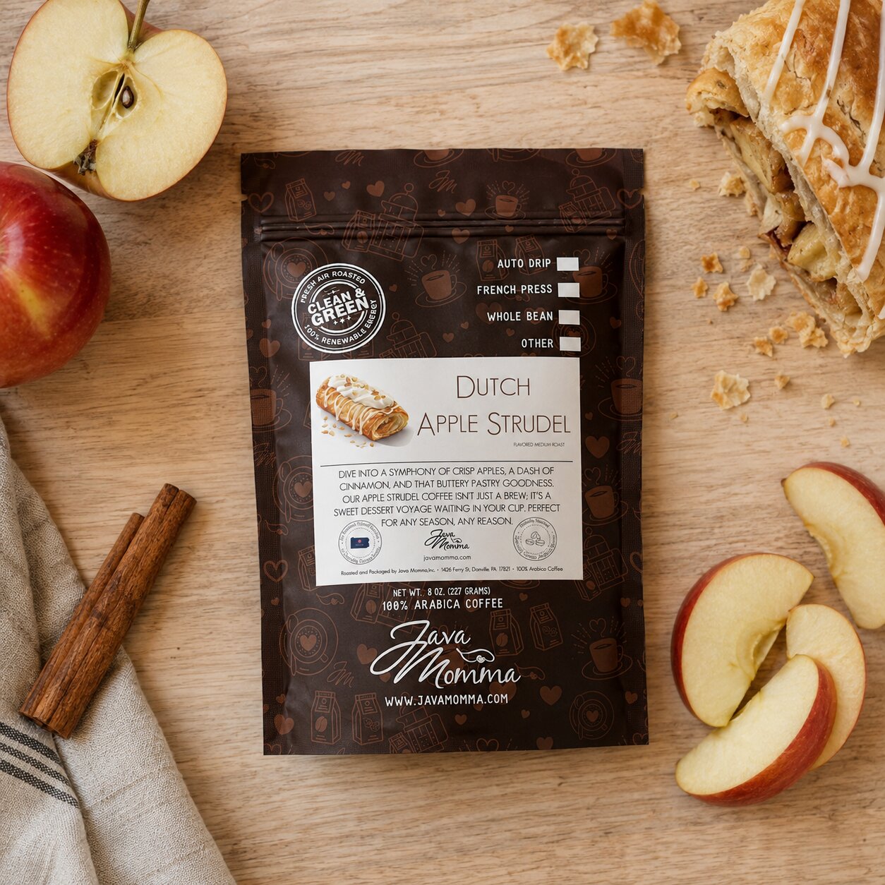 A bag of Java Momma Dutch Apple Strudel coffee sits on a wooden table surrounded by apple slices, a cinnamon stick, a pastry, and a napkin. The package is dark brown with a white label and features the Java Momma brand logo.