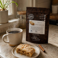 Thumbnail for A bag of Java Momme Dutch Apple Strudel coffee beans stands on a kitchen counter beside a cup of black coffee, a plate with a pastry, and a cinnamon stick. The package is dark brown with a white label and visible Java Momme branding.