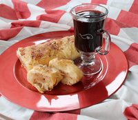 Thumbnail for A red plate holding a golden-brown baked pastry and a clear glass mug filled with a dark beverage, set on a red-and-white checkered fabric background.