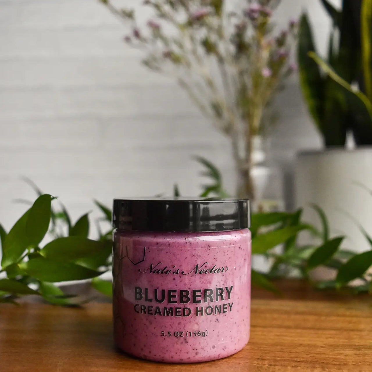 A jar of Nate’s Nectar Blueberry Creamed Honey 💙 – A Berry-licious Spread You’ll Crave sits on a wooden surface among green leaves, with flowers and a white brick wall in the background highlighting its rich blueberry color.