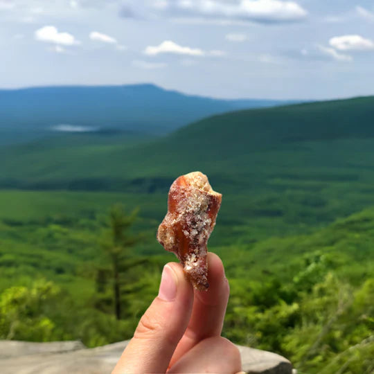 A hand holds up a small, reddish rock with white crystals—just like Sugar + Spice Pretzel Bites: sweet, salty, and straight-up craveable—against lush green hills and a distant blue mountain under a partly cloudy sky.