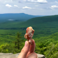 Thumbnail for A hand holds up a small, reddish rock with white crystals—just like Sugar + Spice Pretzel Bites: sweet, salty, and straight-up craveable—against lush green hills and a distant blue mountain under a partly cloudy sky.