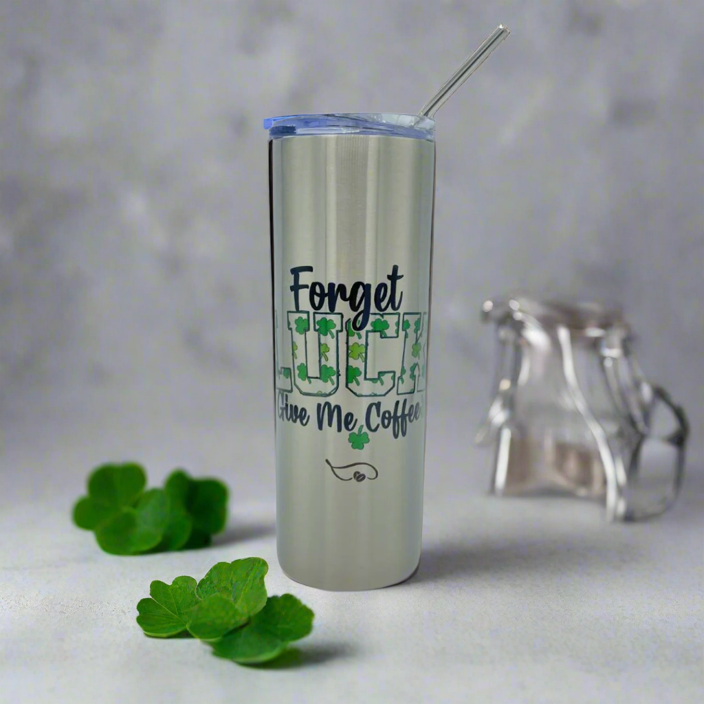 A sleek 20oz stainless steel tumbler with the phrase "Forget Luck, Give Me Coffee" stands upright with a clear lid and straw. The word "LUCK" is decorated with green shamrocks, adding a festive St. Patrick’s Day touch. Surrounded by fresh shamrocks and set against a neutral background, this tumbler is perfect for coffee lovers who believe caffeine is better than luck! 