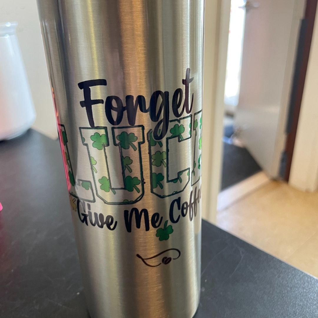 A real-life shot of the Forget Luck, Give Me Coffee tumbler on a dark countertop, with a background showing an open door. The bold lettering and shamrock details pop against the brushed stainless steel, making this the ultimate go-to cup for coffee lovers who run on caffeine—not luck!