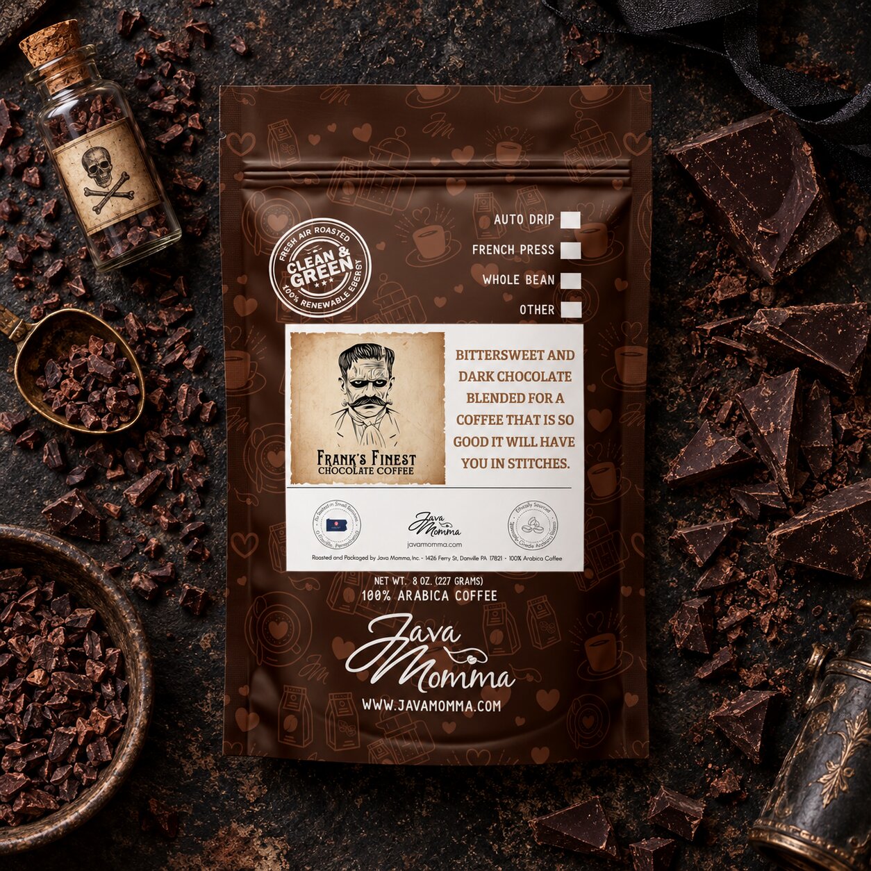 A brown coffee pouch labeled 'Java Momma' and 'Frank & Finest Chocolate Coffee' is photographed flat on a dark textured surface with chocolate pieces, a small labeled glass bottle, and coffee beans around it. No person or animal is present, so this is Product/Packaging rather than Lifestyle.