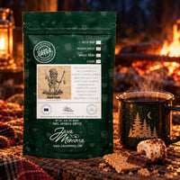 Thumbnail for A bag of Java Momma Ghoul Scout coffee stands next to a steaming dark green enamel mug with a gold tree design, set on a rustic table with marshmallows and a warm fire in the background.
