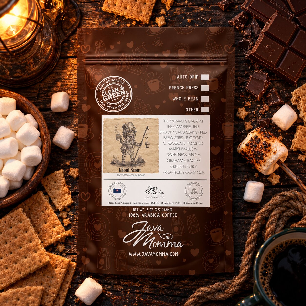 A bag of Java Momma Ghoul Scout
 coffee beans is displayed on a rustic wooden table surrounded by themed props including marshmallows, chocolate, graham crackers, and a cup of coffee.