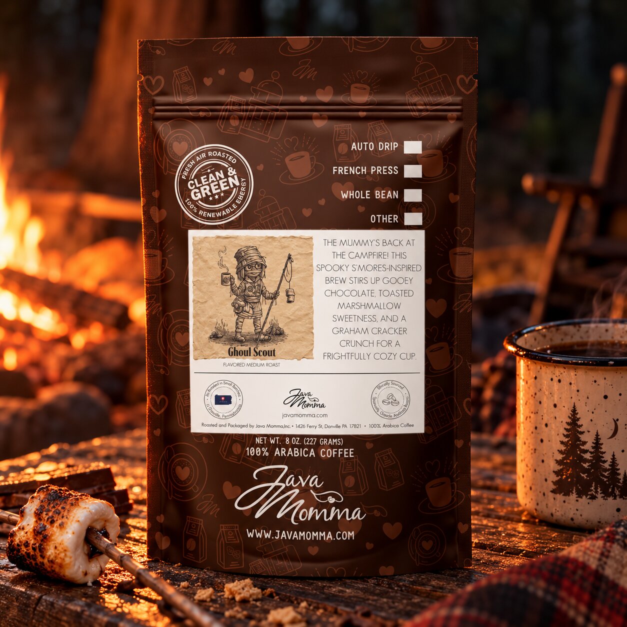 A bag of Java Momma Ghoul Scout coffee stands on a rustic wooden surface in front of a campfire, with a roasted marshmallow on a stick and a speckled mug nearby. The package is dark brown with white and beige printed graphics and text.