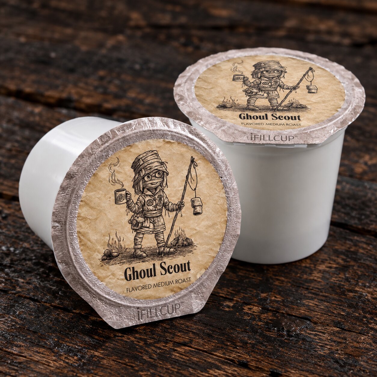Two white iFILL CUP coffee pods with 'Ghoul Scout' flavored medium roast lids, displayed on a rustic dark wood background.