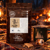 Thumbnail for A bag of Java Momma Clean Green Coffee sits on a campfire with a steaming mug, marshmallow, and blanket nearby. The package is dark brown with white text and graphics, and the background shows warm orange flames and embers.