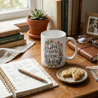 Thumbnail for Mug with 'Give Yourself Time to Bloom' text on a desk with books, a plant, and a plate of cookies.