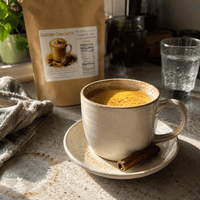 Thumbnail for A ceramic mug filled with golden chai latte sits on a saucer with a cinnamon stick, with a kraft pouch labeled Golden Chai Latte in the background on a kitchen counter.