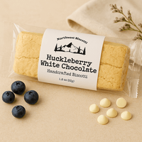 Thumbnail for A Huckleberry White Chocolate Biscotti – Sweet, Tart & Soft-Bite Perfect rests on a beige surface, surrounded by fresh blueberries, white chocolate chips, and a floral sprig for an artful touch.
