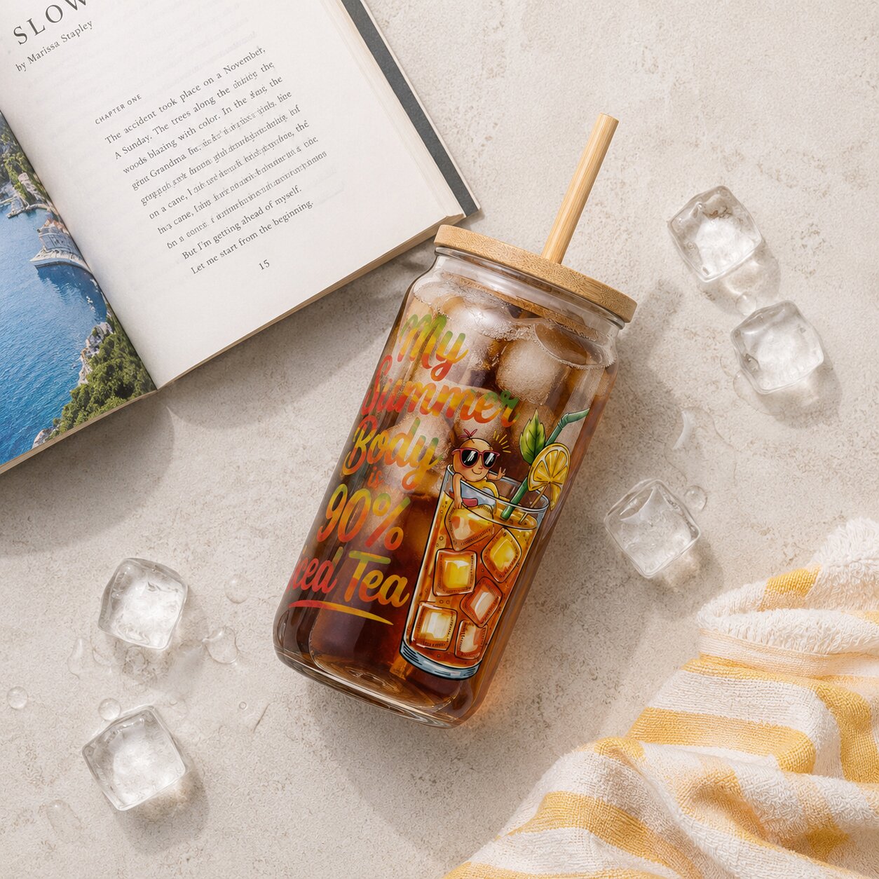 Jar of iced tea with a straw, surrounded by ice cubes and an open book on a light surface.