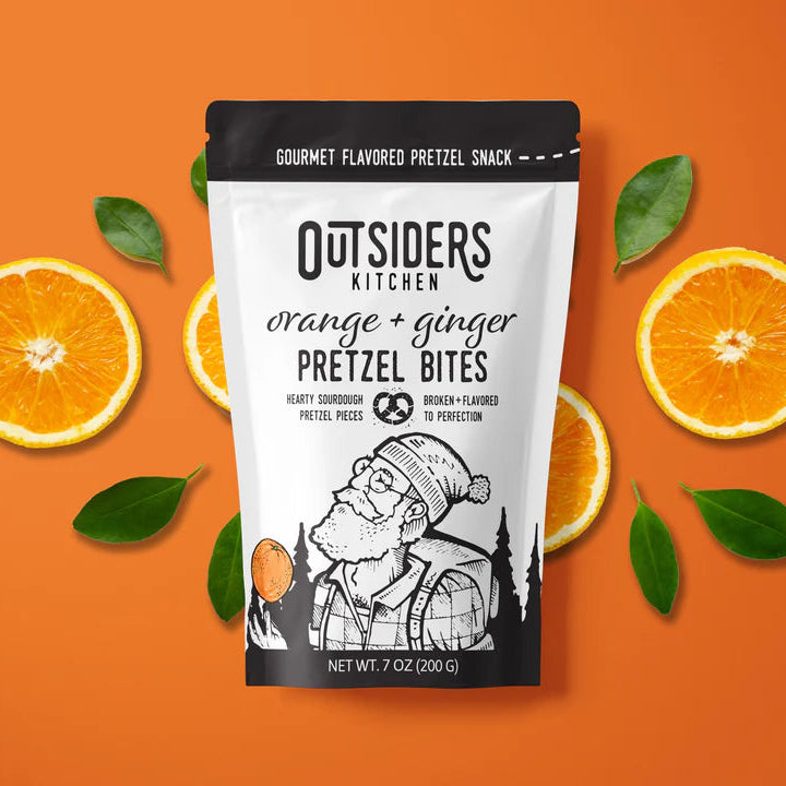 A bag of Orange + Ginger Pretzel Bites – Zesty, Creamy, and Boldly Delicious 🍊🔥 sits on an orange background with citrus accents. The packaging shows an illustrated bearded man in winter gear.