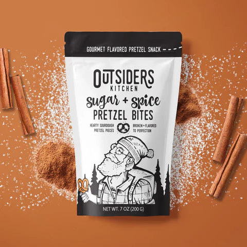 A bag of Sugar + Spice Pretzel Bites – Sweet, Salty, and Straight-Up Craveable 🍬🔥 sits on a brown surface with sugar, cinnamon powder, and sticks. Made in small batches, the packaging features an illustrated bearded man holding a pretzel.