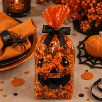 Thumbnail for A clear cellophane bag filled with orange and black popcorn, tied with a black ribbon and decorated with a jack-o'-lantern face, set on a Halloween-themed table with pumpkins, spiderwebs, and candles.