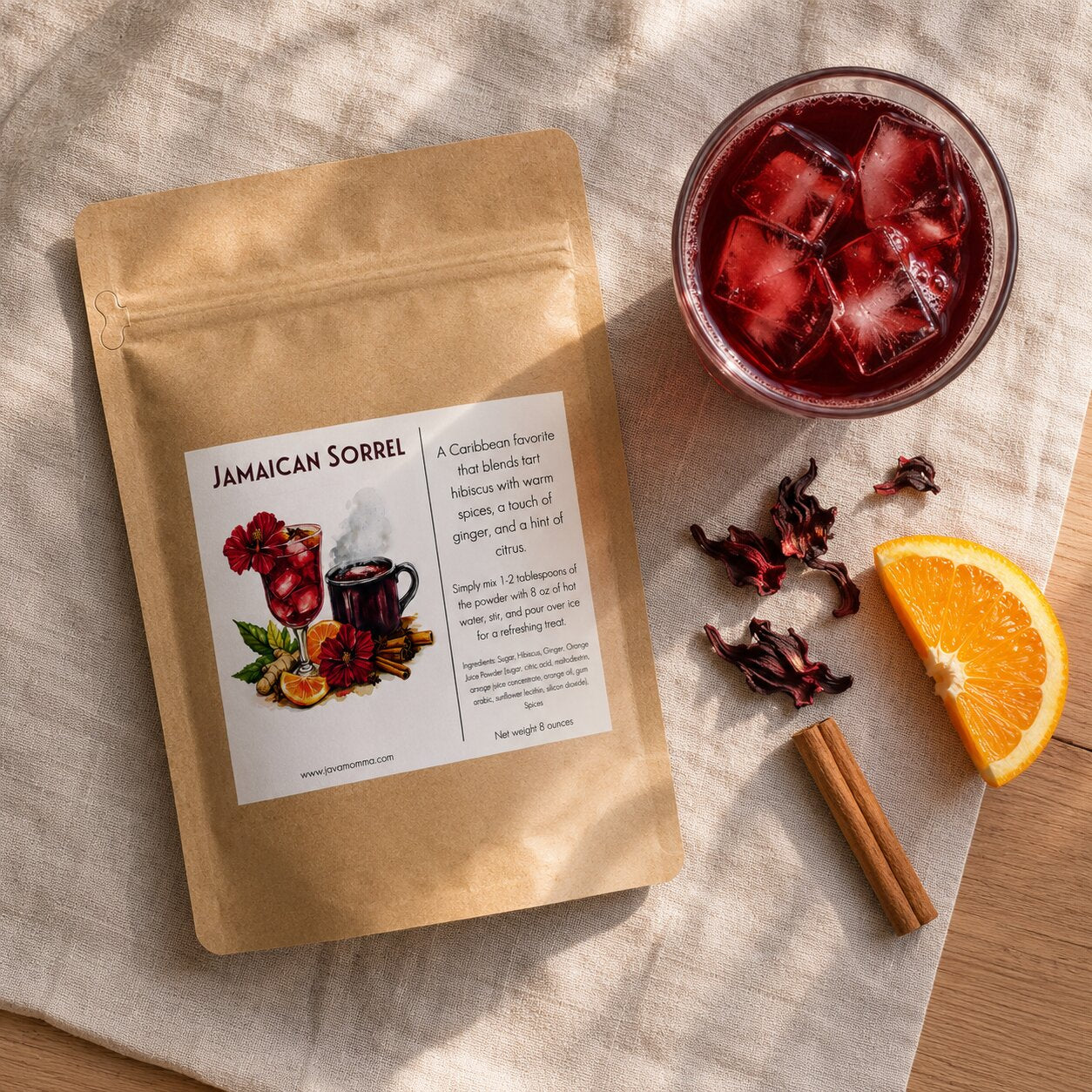 A kraft paper pouch labeled 'Jamaican Sorrel' is arranged on a beige fabric surface with a glass of dark red iced drink, dried hibiscus flowers, a citrus slice, and a cinnamon stick nearby.