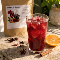 Thumbnail for A glass of iced Jamaican sorrel drink with ice cubes and visible bubbles, set on a stone surface beside a sliced orange, cinnamon stick, dried flowers, and a kraft pouch labeled Jamaican Sorrel. A real tabletop environment and plants are visible, so this is Lifestyle rather than Product/Packaging.